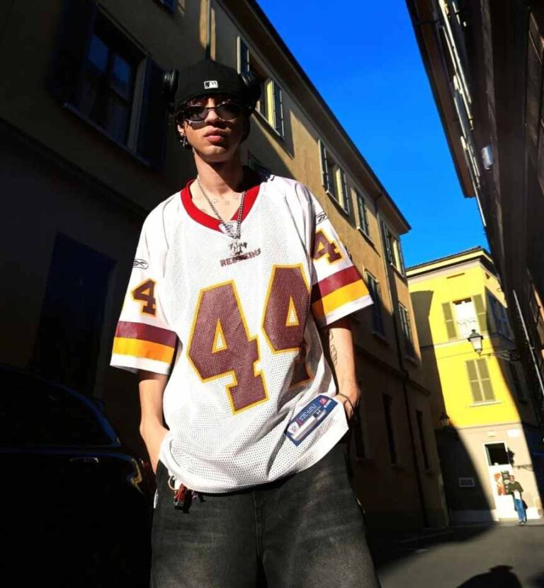 guy wearing an oversized white sports jersey with red and yellow accents paired with baggy black jeans