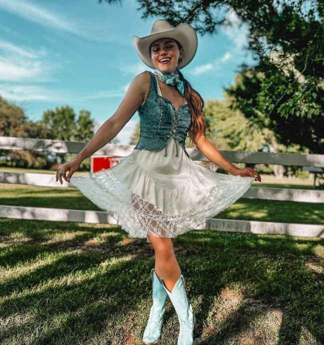 Cara wearing a a blue corset-style denim vest over a white breezy dress with cowboy boots and hat