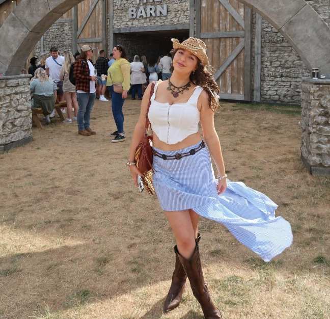 Lydia wearing a white bustier crop top with a blue and white asymmetrical skirt and brown cowboy boots