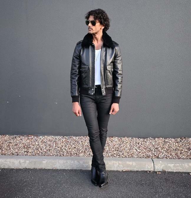 Guy wearing a black leather bomber jacket over a white tank top and blakck skinny jeans