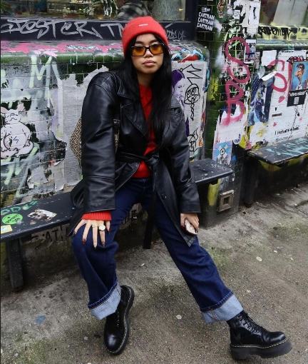 girl wearing a belted black leather coat over A bright red sweater paired with cuffed dark-wash jeans and doc martens