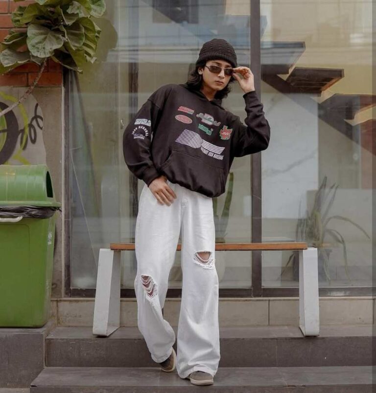 guy wearing A brown oversized hoodie featuring colorful graphic patches and prints is paired with baggy white jeans with distressed details