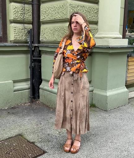 woman wearing A vibrant wrap blouse featuring orange, yellow, and brown floral patterns with a tan suede button-down midi skirt